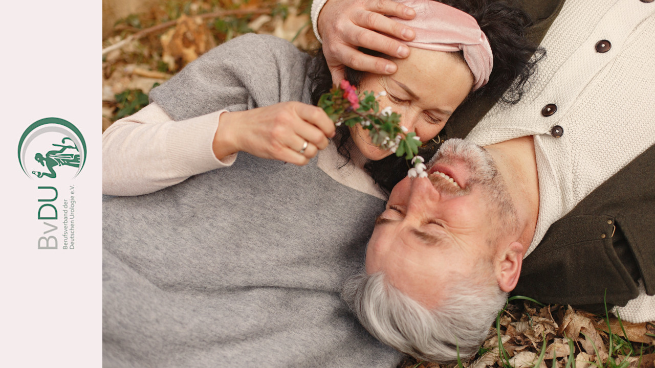 Ein älteres paar liegt auf dem Herbstlaub auf dem Rücken. Sie spielt liebevoll mit einem Blütenstengel an seiner Nase herum.