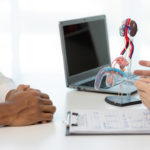 Arzt im Gespräch mit einem Patienten. Im Hintergrund ein Laptop und ein anatomisches Modell