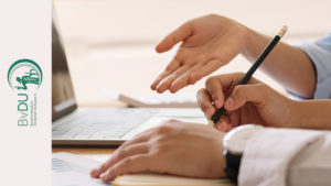 Zwei Personen diskutieren vor einem Laptop. Eine Person schreibt mit, die andere zeigt mit offener Hand auf eine Fragestellung