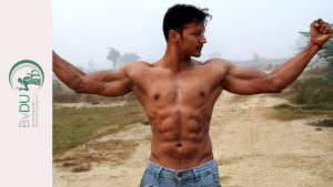 Ein Mann der mit nacktem Oberkörper und weit von sich gestreckten Armen seine Muskeln zur Schau stellt.